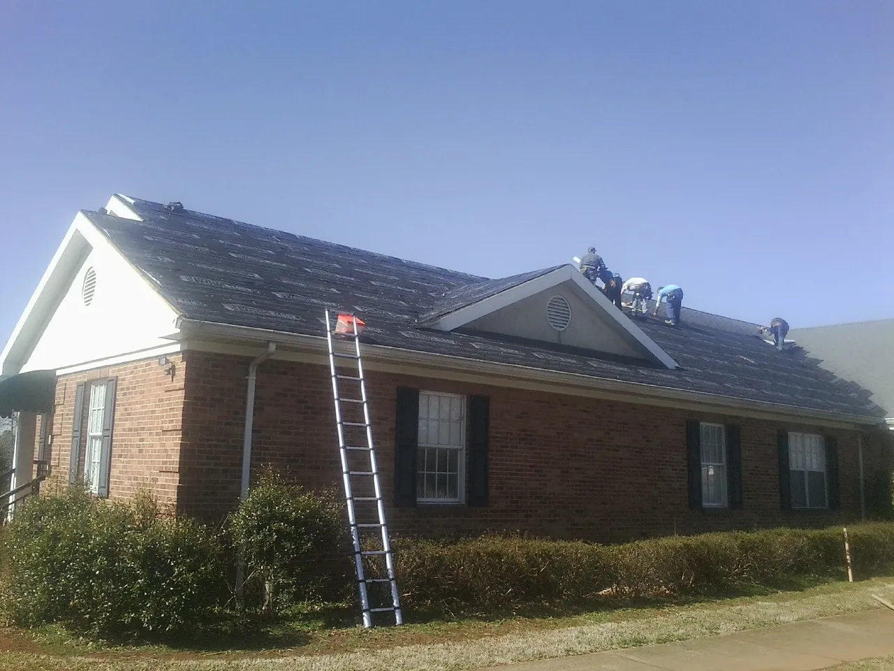 Crew tearing shingles off a brick ranch roof with a ladder set — Emergency Roof Repair work by our crew in Rockville